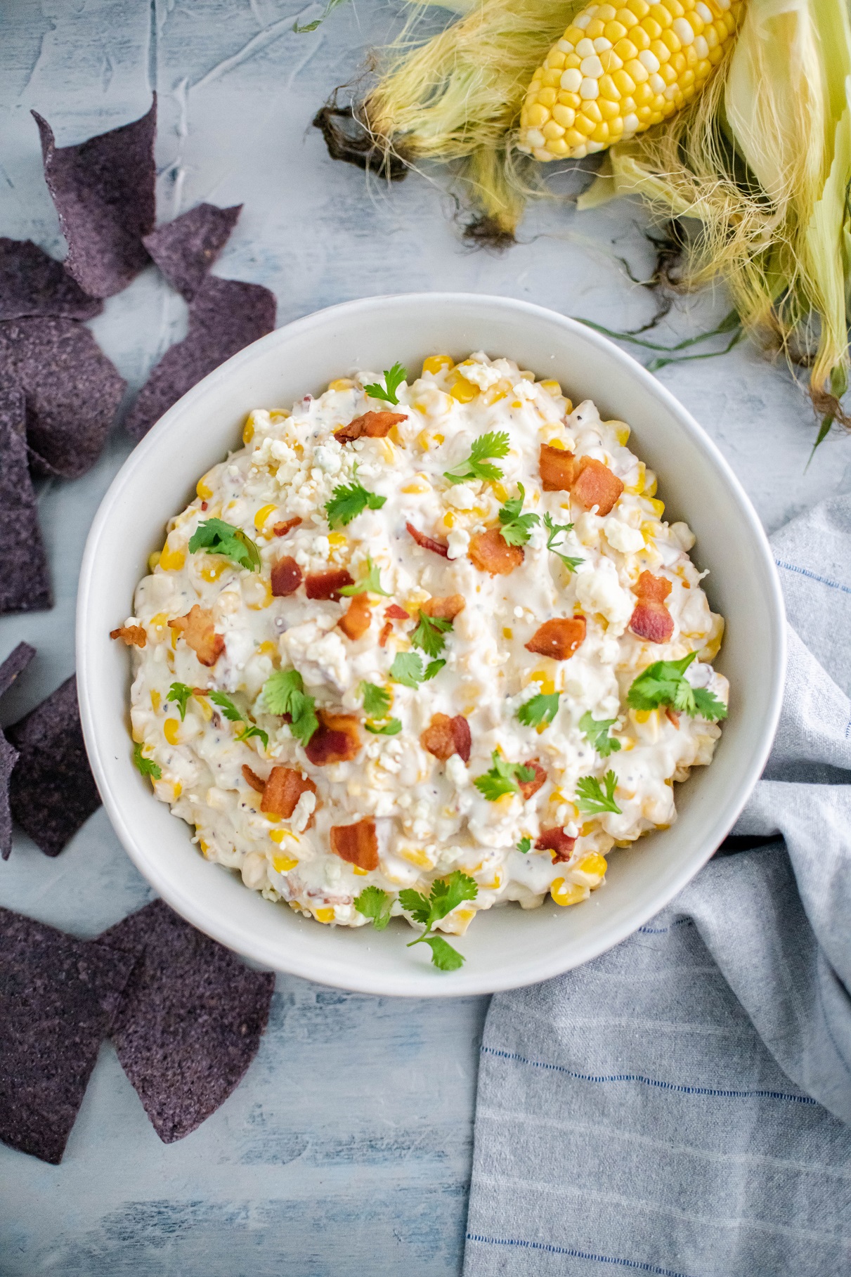 White bowl filled with hot corn dip, topped with crumbled bacon and cilantro. Husked corn in upper right corner and blue corn tortilla chips sprinkled around.