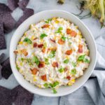 White bowl filled with hot corn dip, topped with crumbled bacon and cilantro. Husked corn in upper right corner and blue corn tortilla chips sprinkled around.