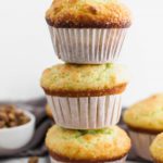 3 large bakery style pistachio muffins stacked on top of each other with bowl of pistachios in upper left corner and more muffins in upper right corner.