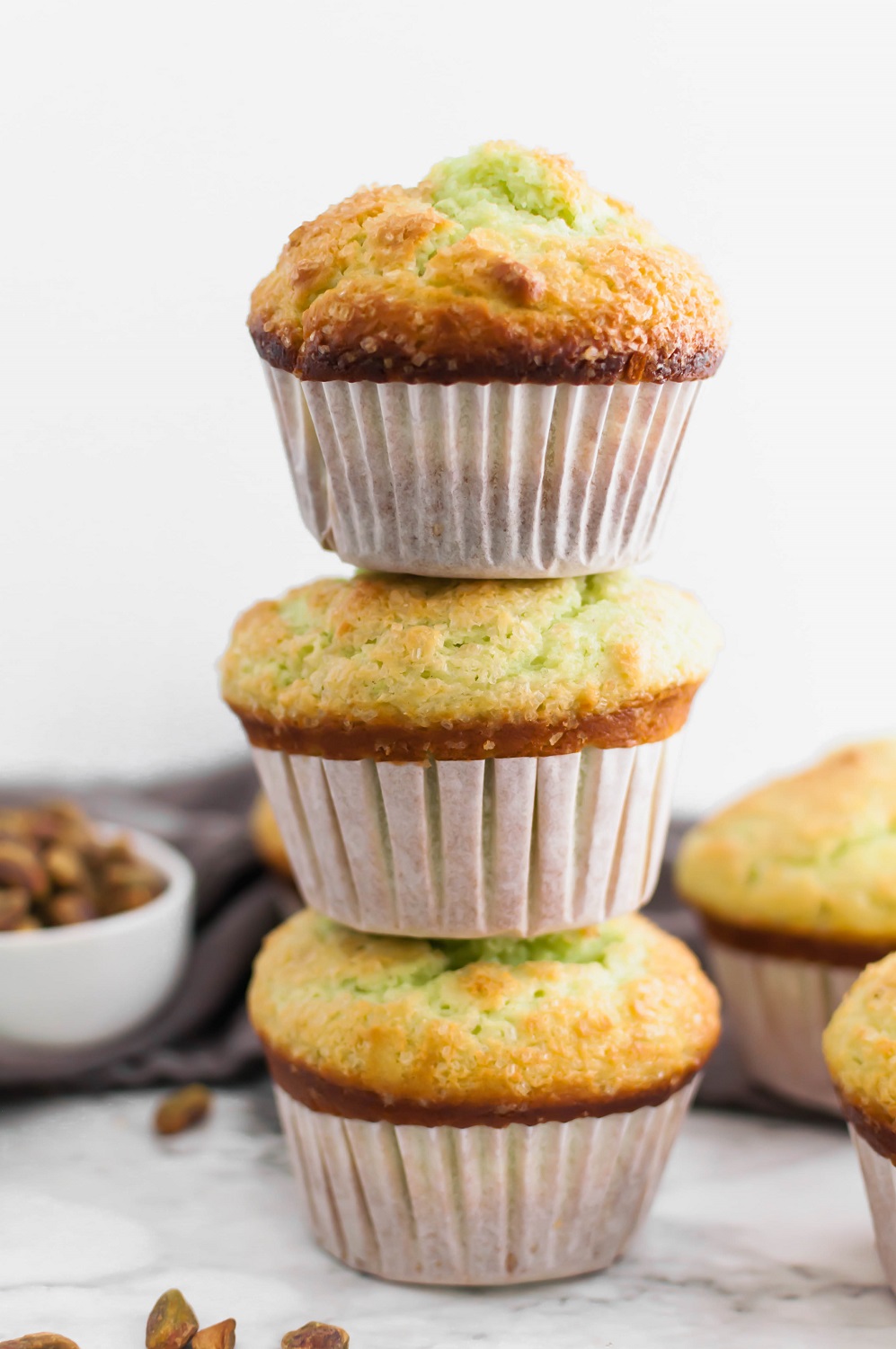 3 large bakery style pistachio muffins stacked on top of each other with bowl of pistachios in upper left corner and more muffins in upper right corner.