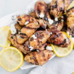 Basket of grilled Greek chicken wings garnished with crumbled feta and thin lemon slices. Gray striped napkin in bottom right corner.