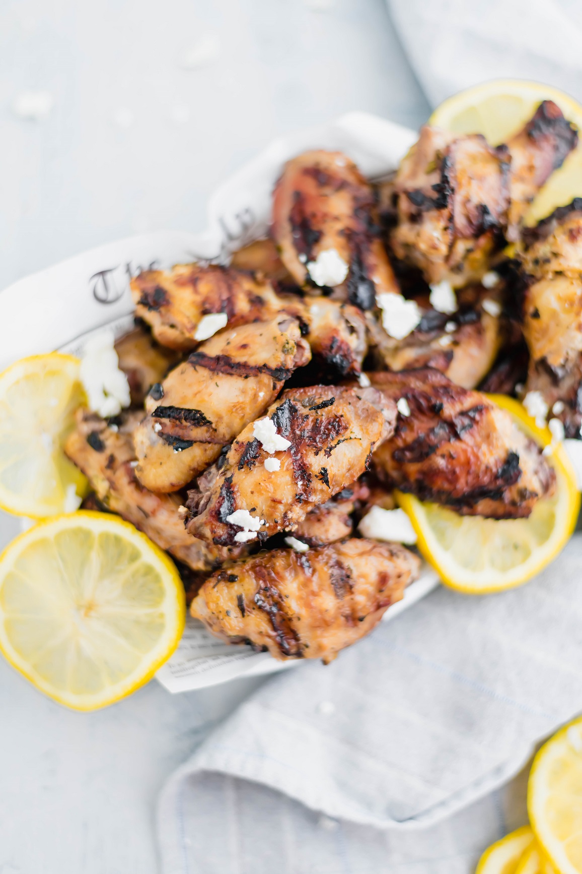 Basket of grilled Greek chicken wings garnished with crumbled feta and thin lemon slices. Gray striped napkin in bottom right corner.