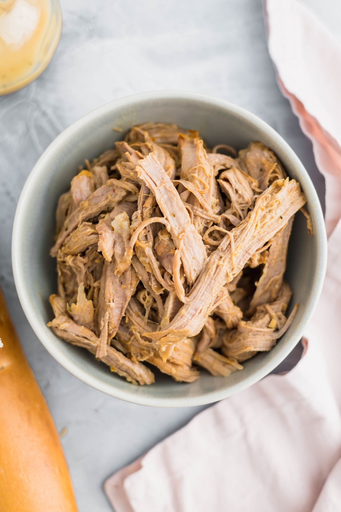 Shredded slow cooker Italian pork in a gray bowl. Bottle of dressing in upper left corner and baguette in lower left corner. Pale pink cloth napkin draped on right side of photo.