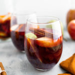 Two glasses of fall sangria with sliced apples, sliced pears, sliced oranges and a cinnamon stick floating in it. Pitcher of sangria in back left corner.