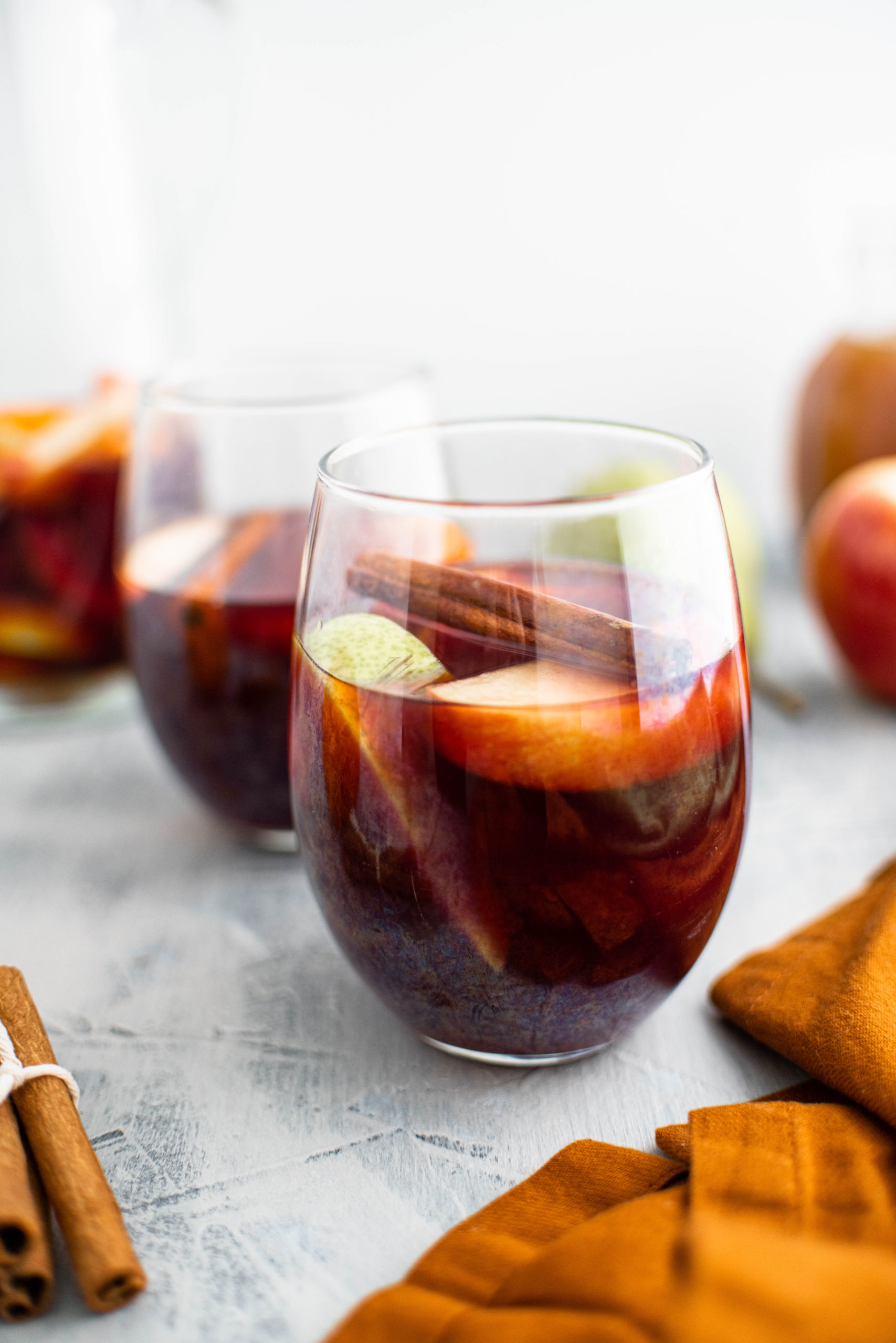 Two glasses of fall sangria with sliced apples, sliced pears, sliced oranges and a cinnamon stick floating in it. Pitcher of sangria in back left corner.