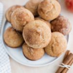 Dozen apple cider muffins piled on a round white plate with a tied bundle of cinnamon sticks in lover right corner and apples and apple cider in background.