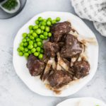 Tender instant pot beef tips over egg noodles with a side of peas on a small white round plate. Another prepared plate peeking in lower right corner.