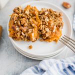 Two scoops of sweet potato crunch on a stack of small round white plates. Fork leaning on the edg of the plate with a blue and white striped cloth napkin in lower right corner.