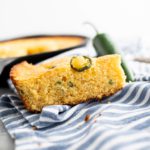 Slice of jalapeno cheddar cornbread on a blue and white striped cloth napkin with cast iron skillet of cornbread in background.