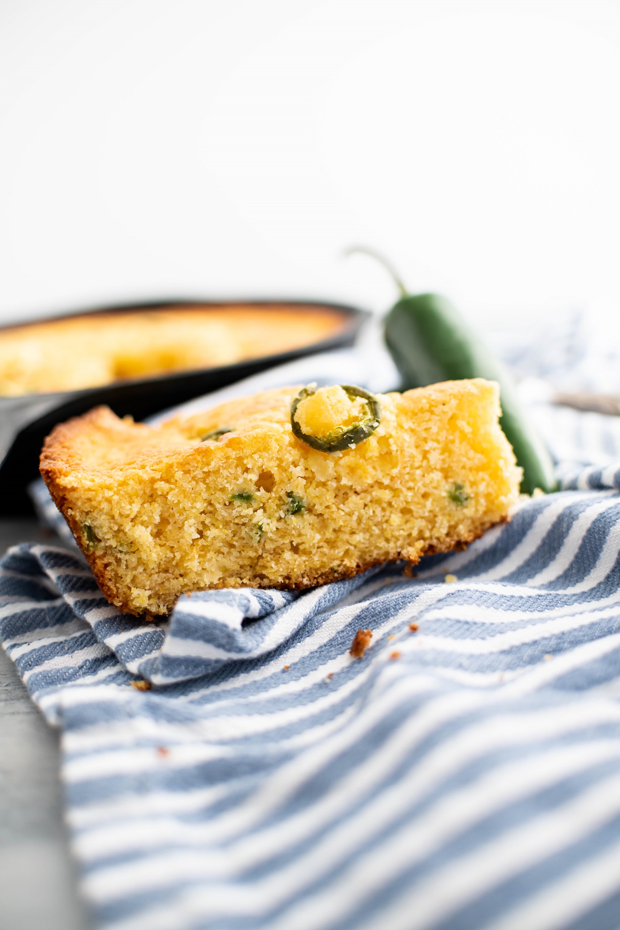 Slice of jalapeno cheddar cornbread on a blue and white striped cloth napkin with cast iron skillet of cornbread in background.