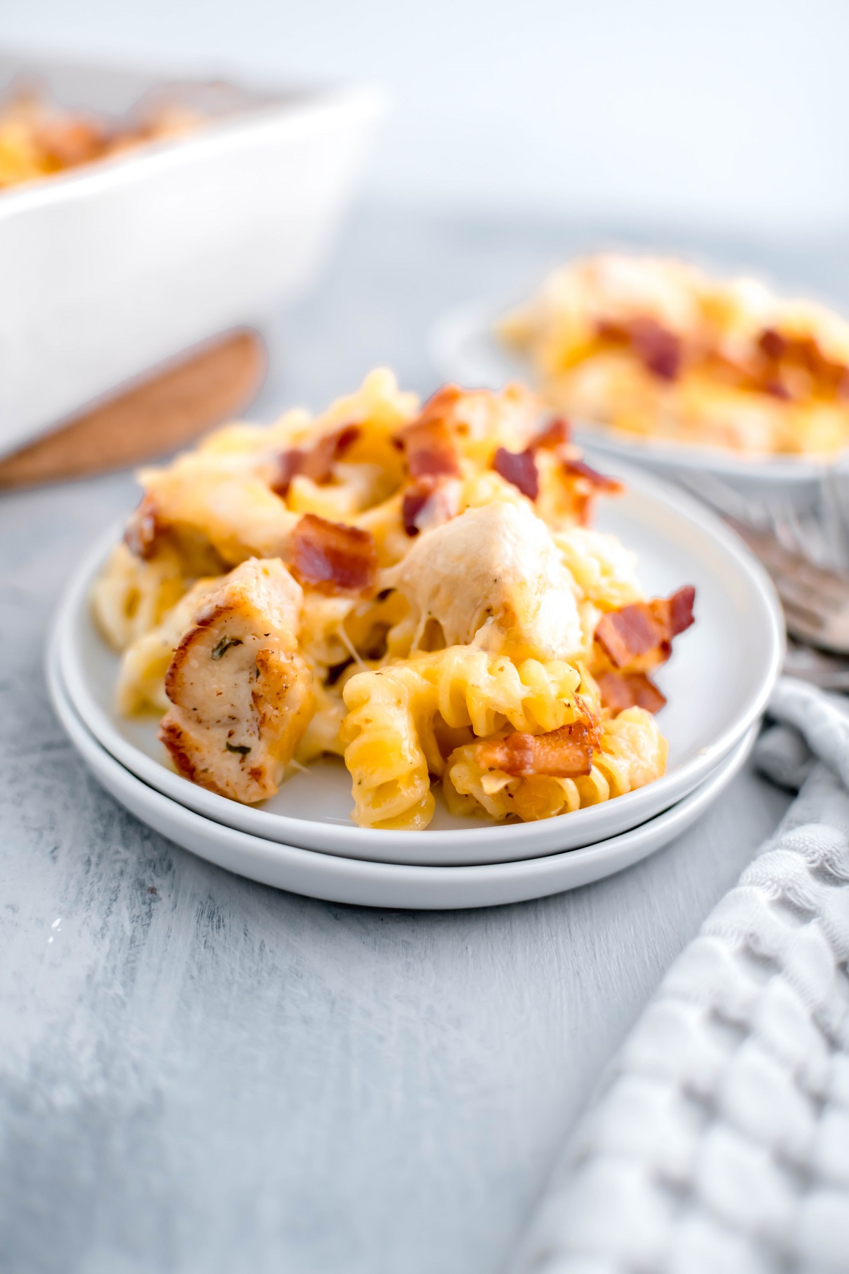 Portion of chicken bacon ranch casserole on a small white round plate with another plate filled in the background. Casserole dish to the left.