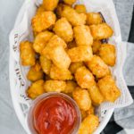 White plastic basket lined with newspaper parchment and filled with crispy tater tots and a small glass bowl of ketchup