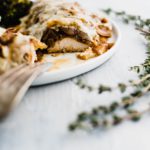 Chicken covered in caramelized onions and melted swiss cheese on a round white plate. Bite cut out of it to show interior.