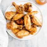 White round plate piled with hot honey wings. Glass jar of honey with a dipper in the background.