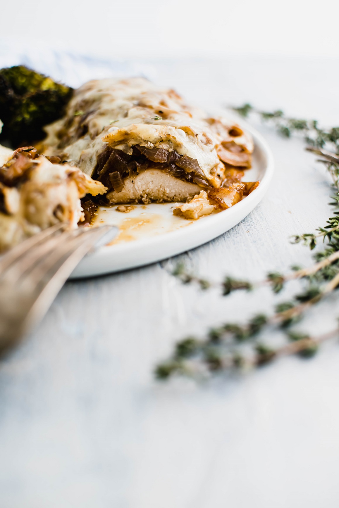 Chicken covered in caramelized onions and melted swiss cheese on a round white plate. Bite cut out of it to show interior.