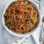 White bowl filled with veggie lo mein. Blue and white striped napkin to the right with a pile of antique forks.