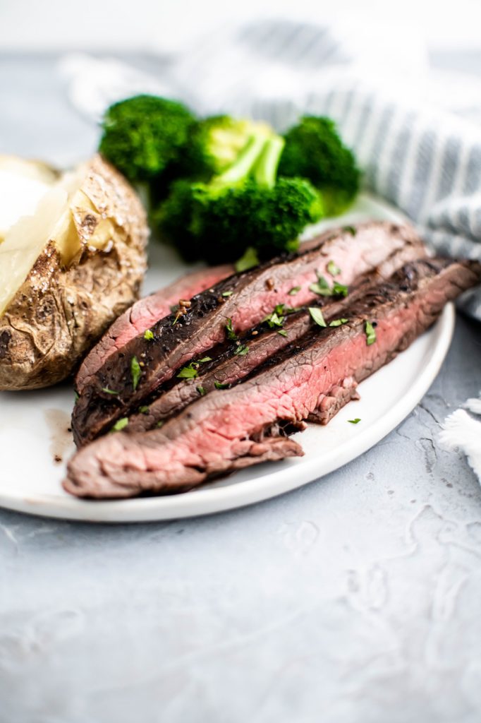 Thin slices of flank steak on a white round plate with a baked potato and steamed broccoli.