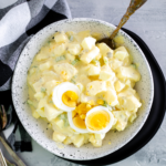 White bowl filled with potato salad. Set on top of a black plate.