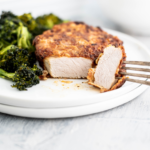 Southern Fried Pork Chop on a round white plate with a bite cut off and on the fork to show the interior of the pork chop. Roasted broccoli on the side.