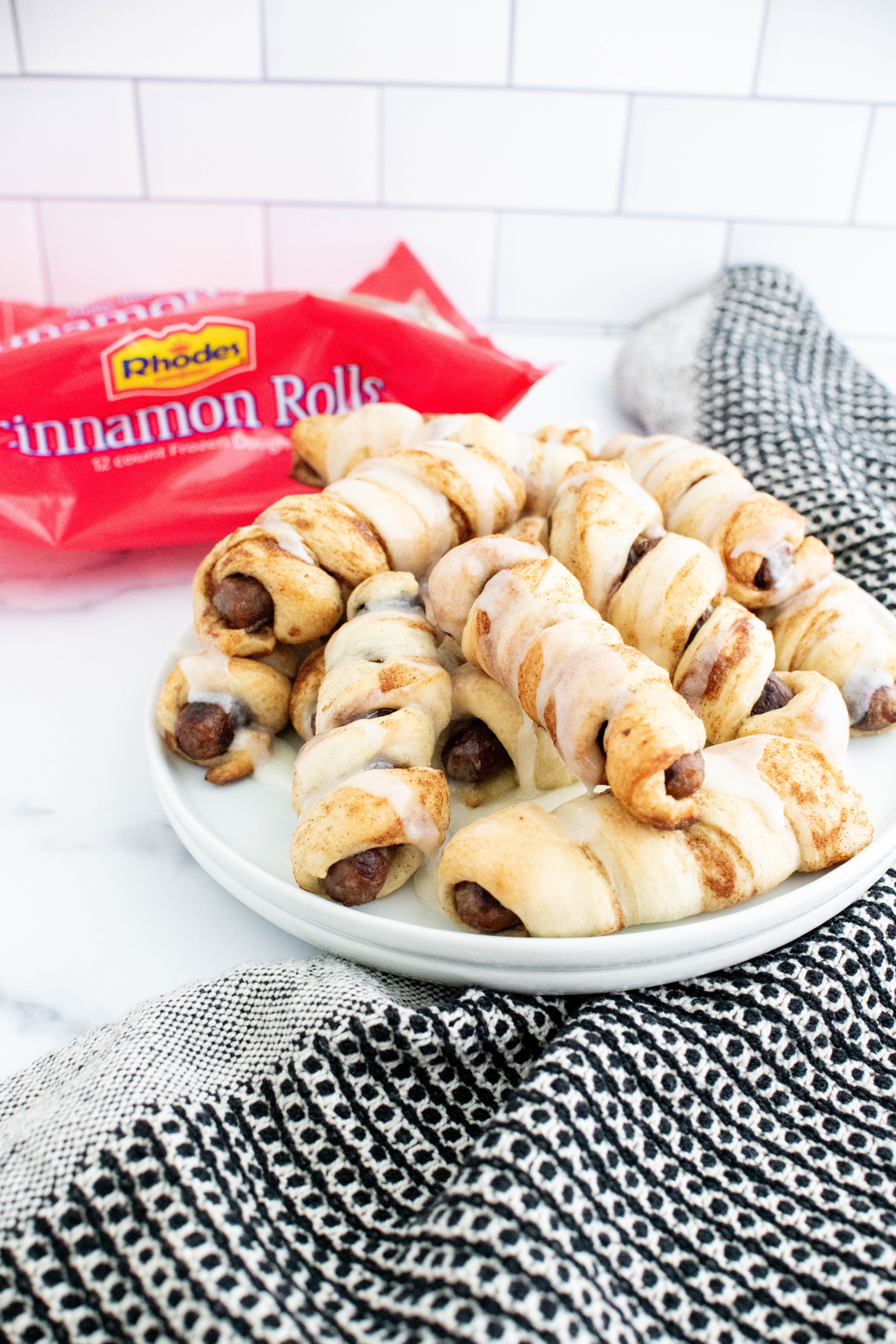 White round plate piled with cinnamon roll pigs in a blanket that are drizzled with icing. Rhodes cinnamon roll package in the background.