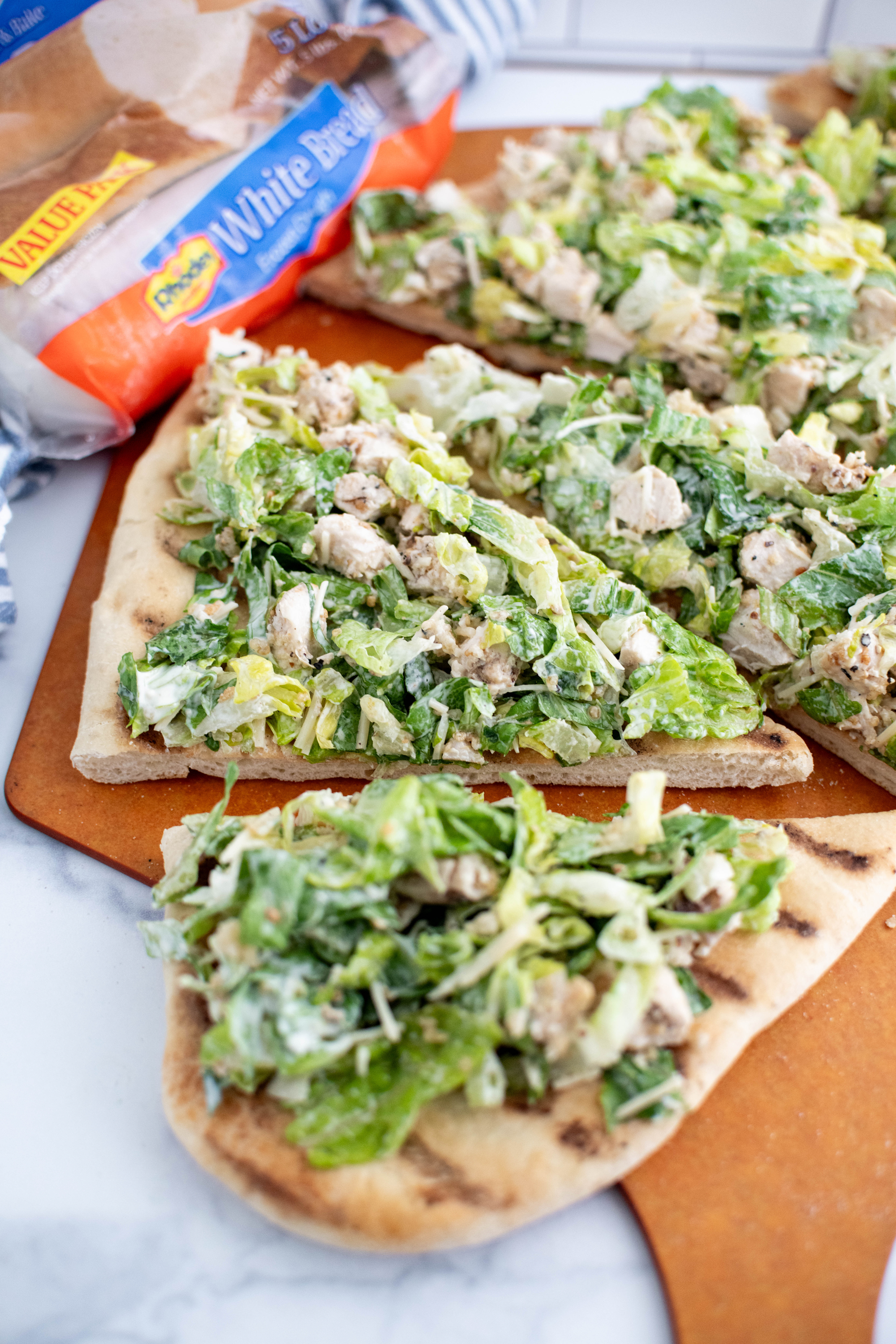 Grilled chicken caesar salad flatbread sliced into triangles on a pizza peel. Package of Rhodes bread dough in the background.