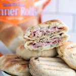 Grilled Cuban Sandwich Pockets in a pile on a roun white plate with one cut in half to show the interior. Bag of Rhodes bread rolls in the background.